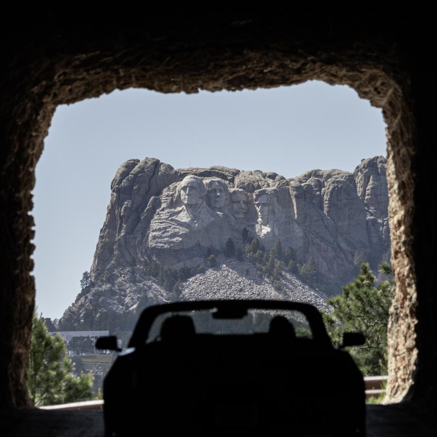 The photo captures Mount Rushmore framed through a tunnel. A silhouetted car is driving through the tunnel with the iconic Mount Rushmore National Monument in the background.